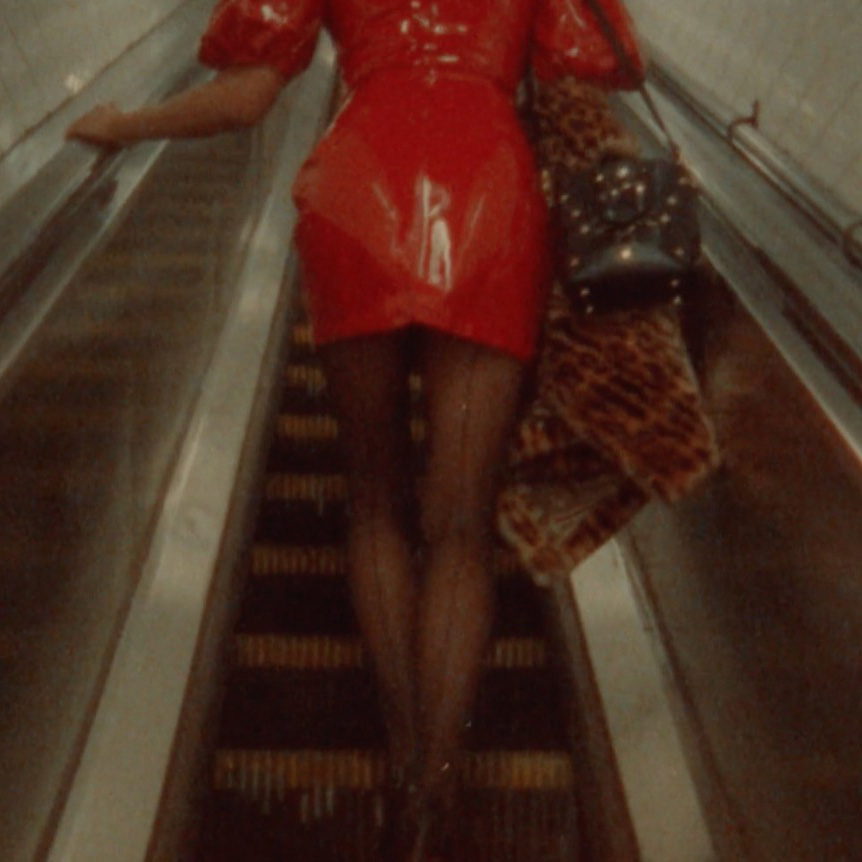 A photograph with a warm, grainy, retro quality shows a woman riding an escalator, her back-seamed stockings leading up to a shiny red vinyl dress, with a furry, animal print coat hanging from her arm, her head cropped from the frame.