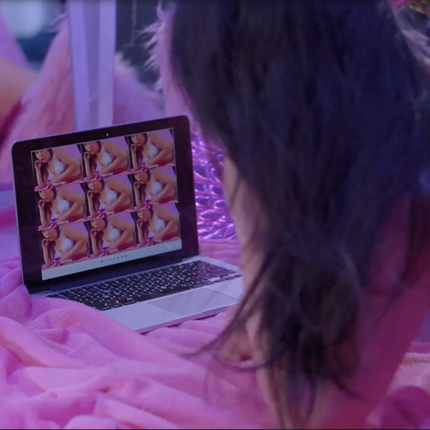 A femme person reclines on pink fabric facing an open laptop, their pose reflected multiple times on its screen.