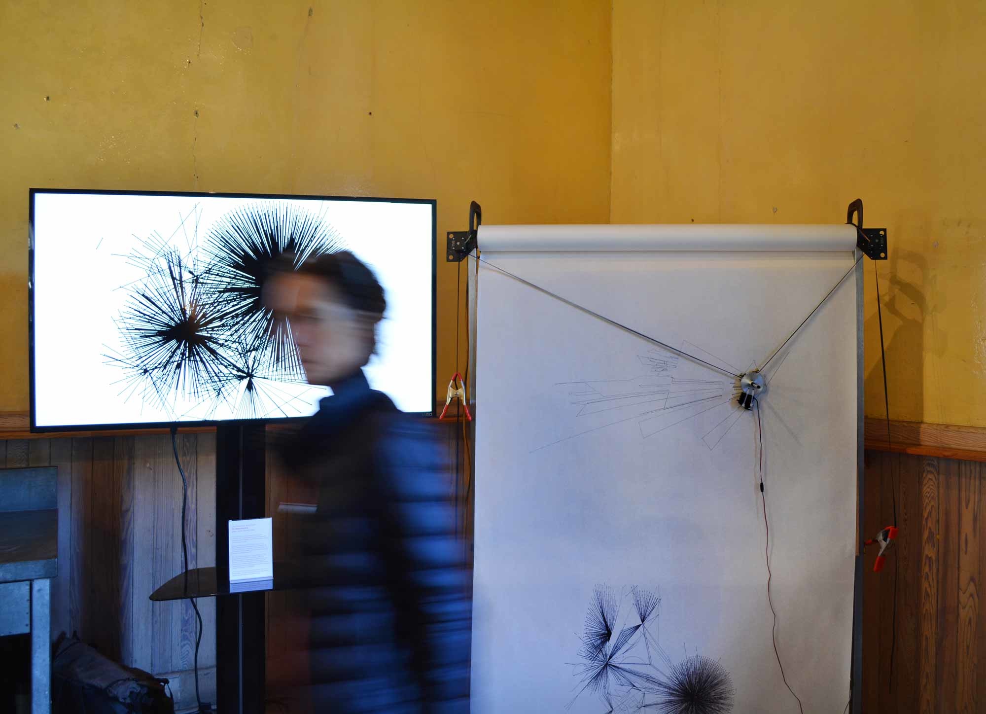 In a warm yellow room, a large video monitor displaying radiating forms stands beside a drawing machine marking a long paper scroll, as a visitor with a motion blurred face walks past.